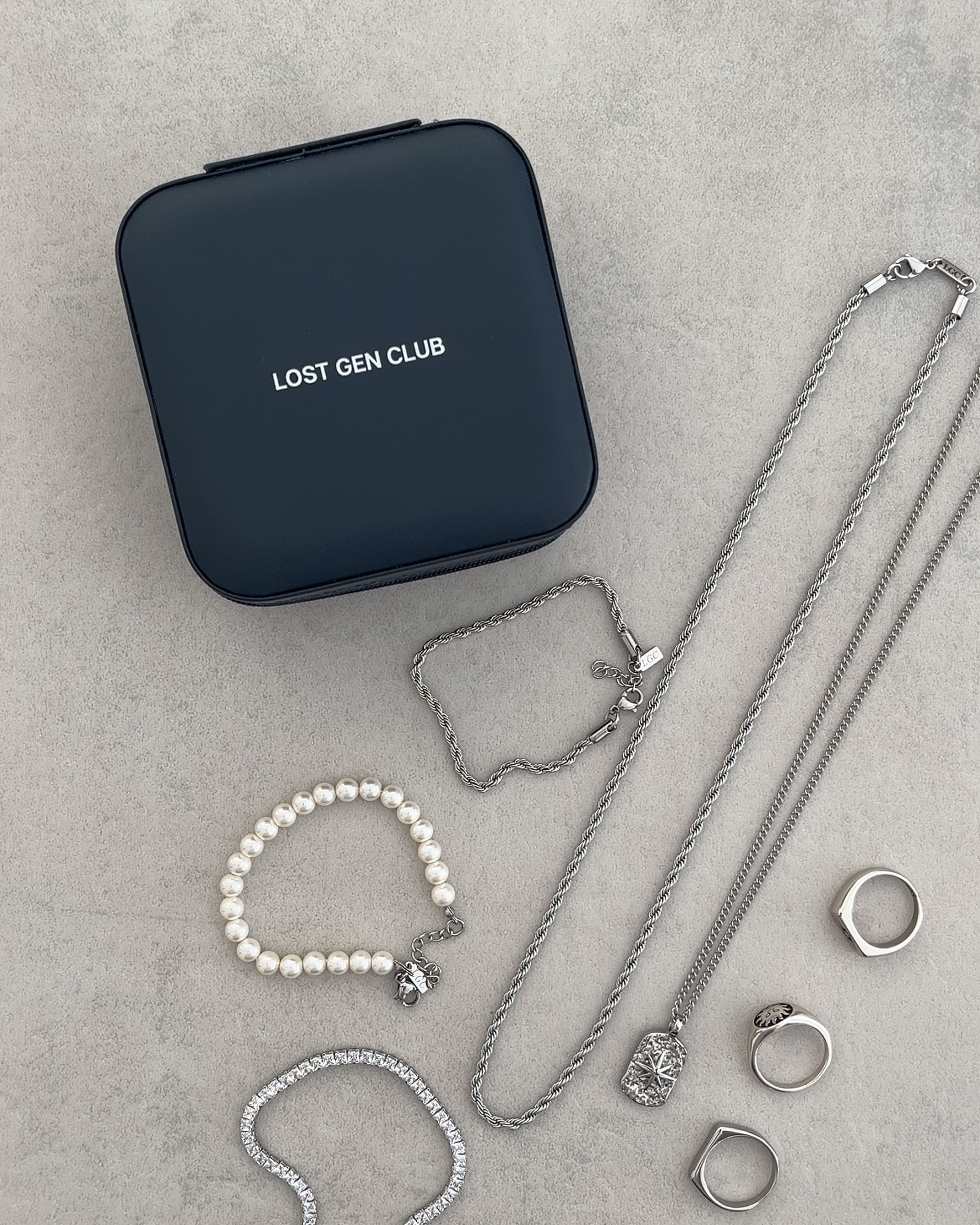 Blue travel jewelry case with LOST GEN CLUB branding surrounded by silver chains and a pearl bracelet.