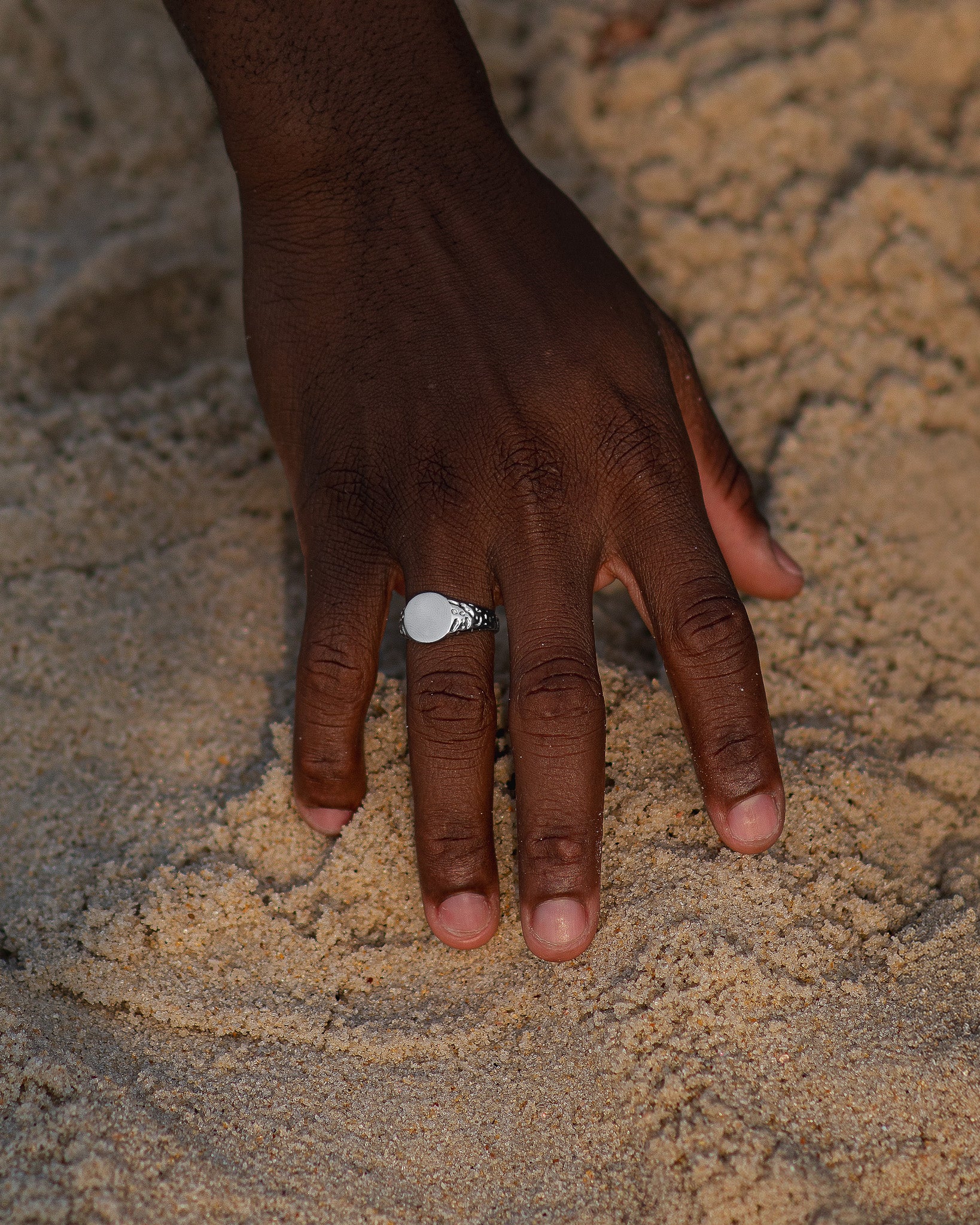 Front view of a silver EDEN ring with a smooth circular face on the wearer's finger against a sandy background.