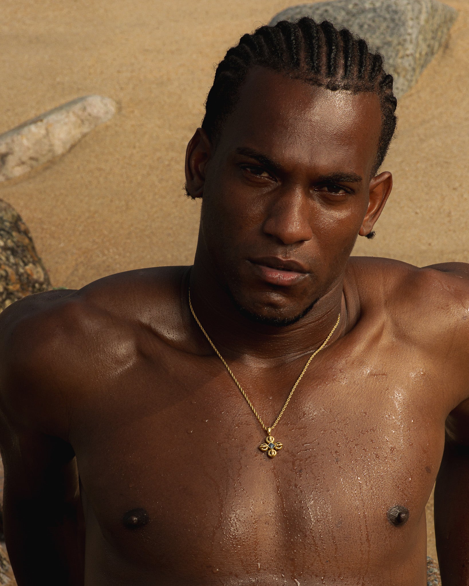 Portrait of a man with braided hair wearing a gold necklace against a sandy outdoor background.