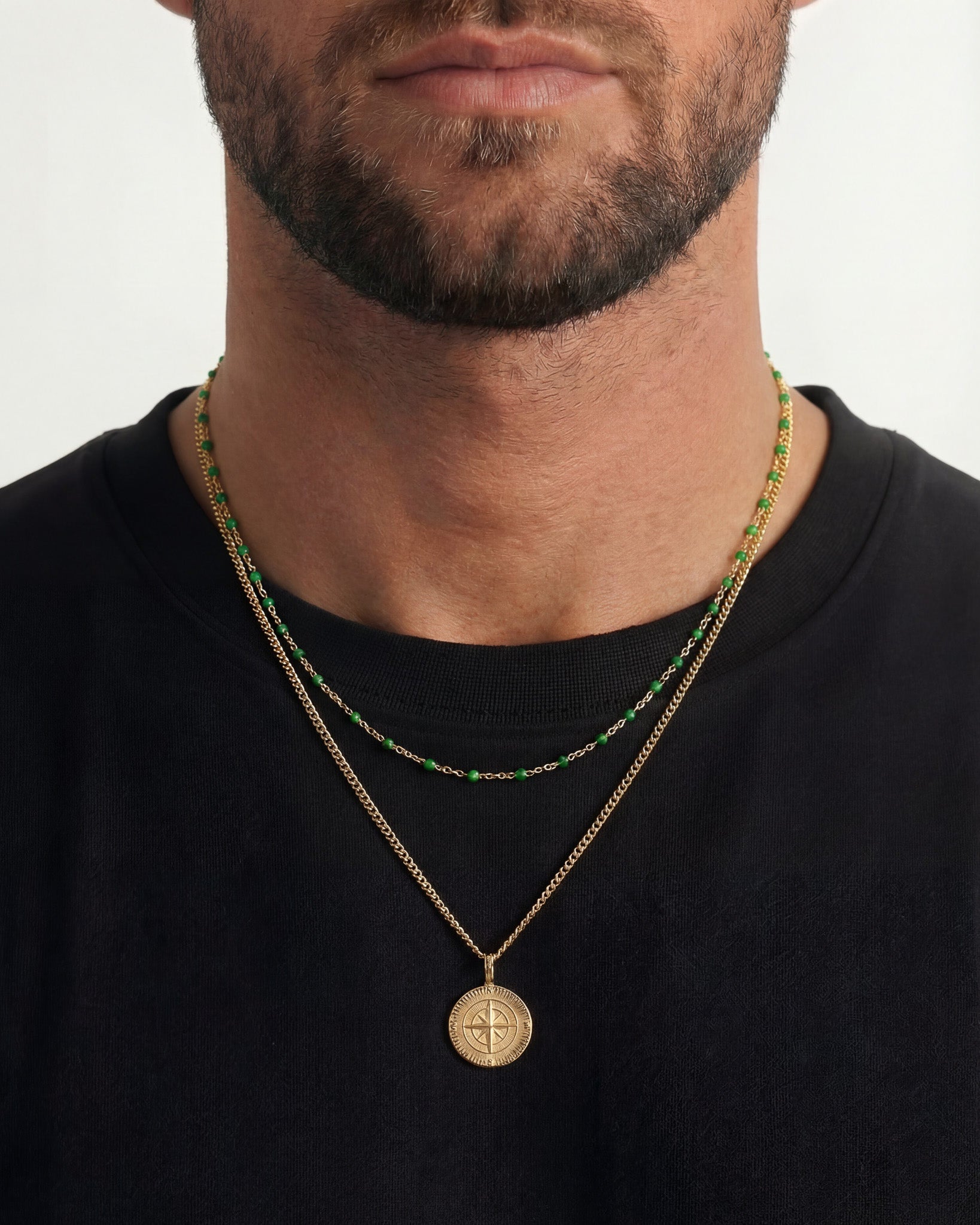 Close up of a man wearing a gold necklace with green beads and a compass pendant resting on the chest