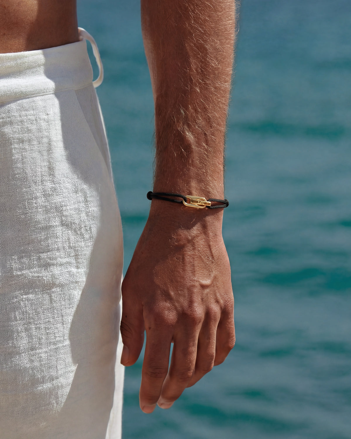 Atlantis gold bracelet with black cord and a gold clasp worn on the wrist by the sea