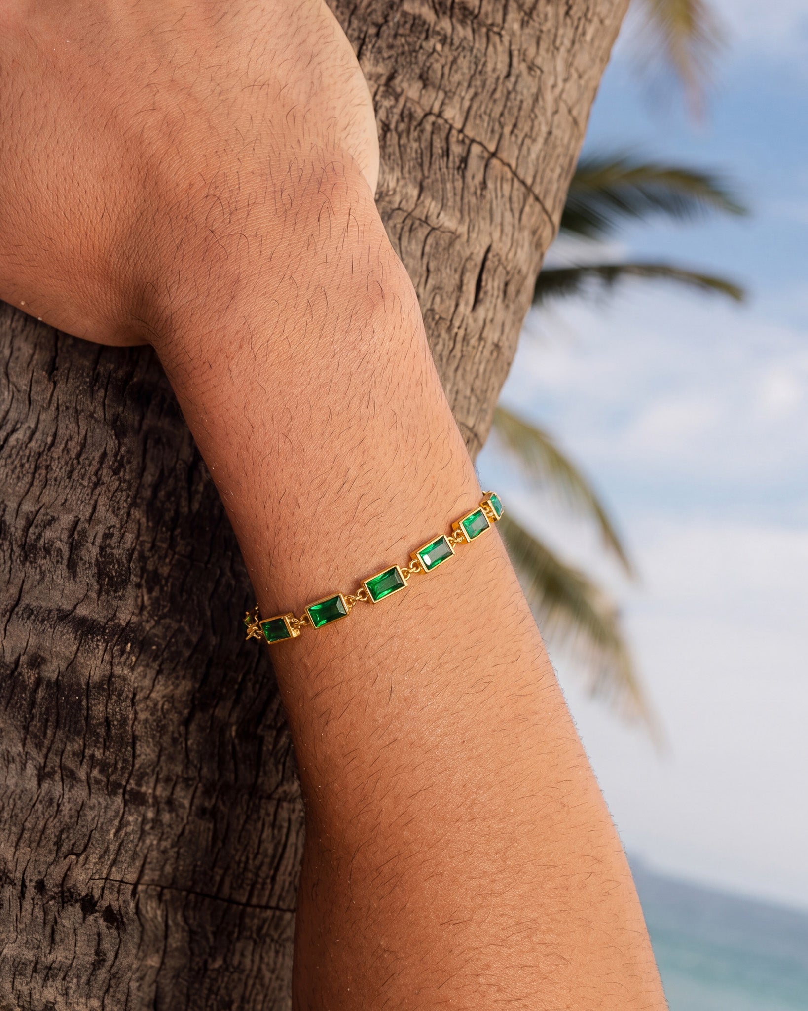 Primer plano de la pulsera OMEN de oro con piedras verde esmeralda en la muñeca, al aire libre junto a un tronco